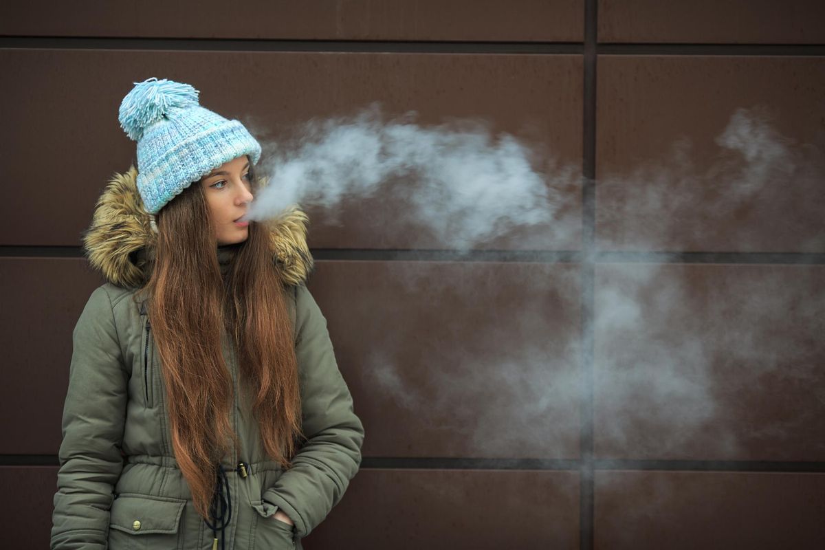 Vape teenager. Young pretty white girl in a cap smoking an electronic cigarette opposite modern brown background on the street in the winter. Bad habit. Close up.