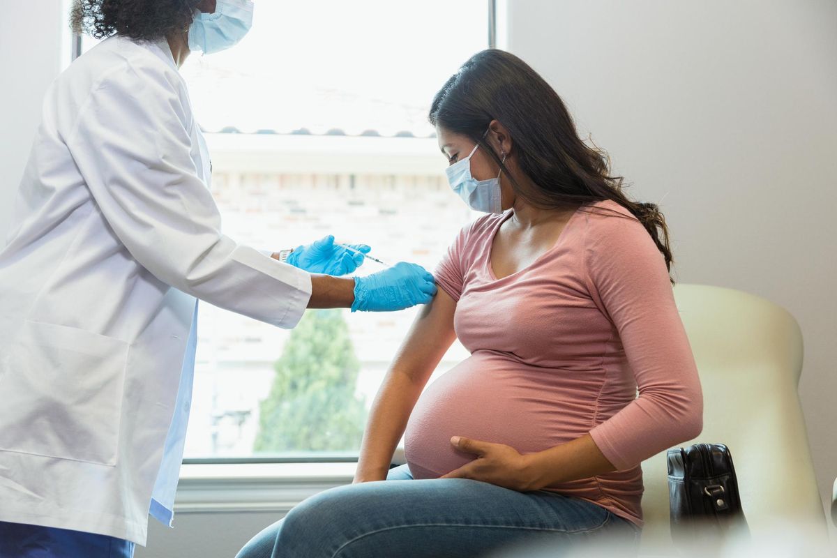 Wearing protective masks because of COVID-19, a mid adult pregnant woman watches as the female technician administers a booster shot.