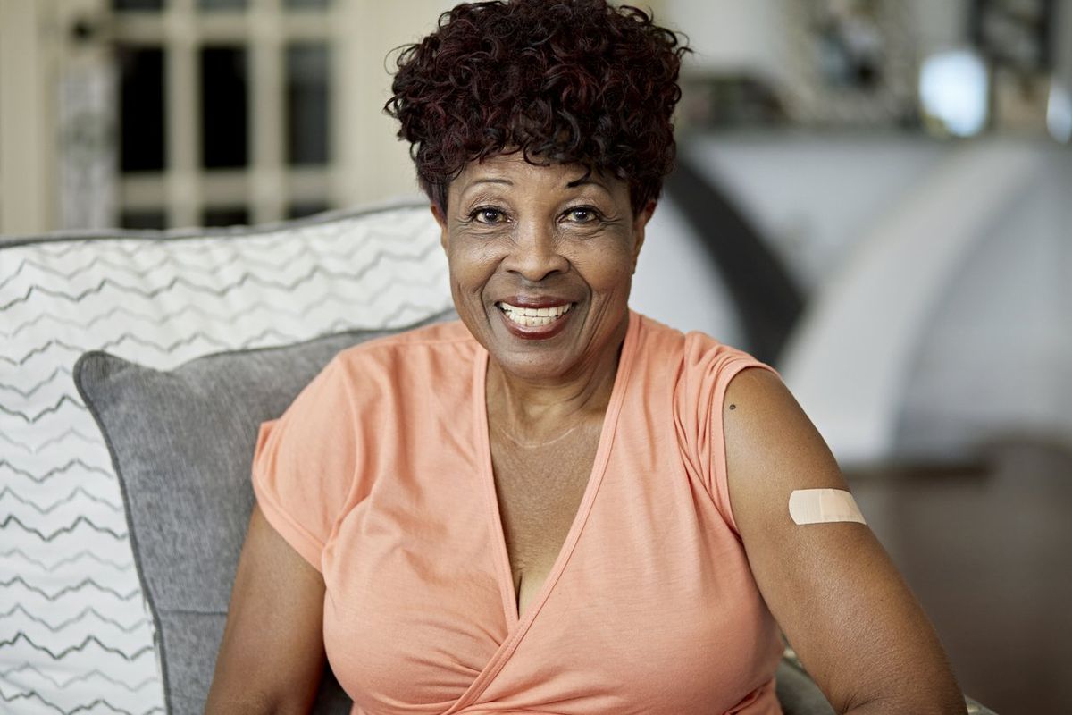 woman in casual clothing with visible adhesive bandage from vaccination sitting on sofa in family home and smiling at camera