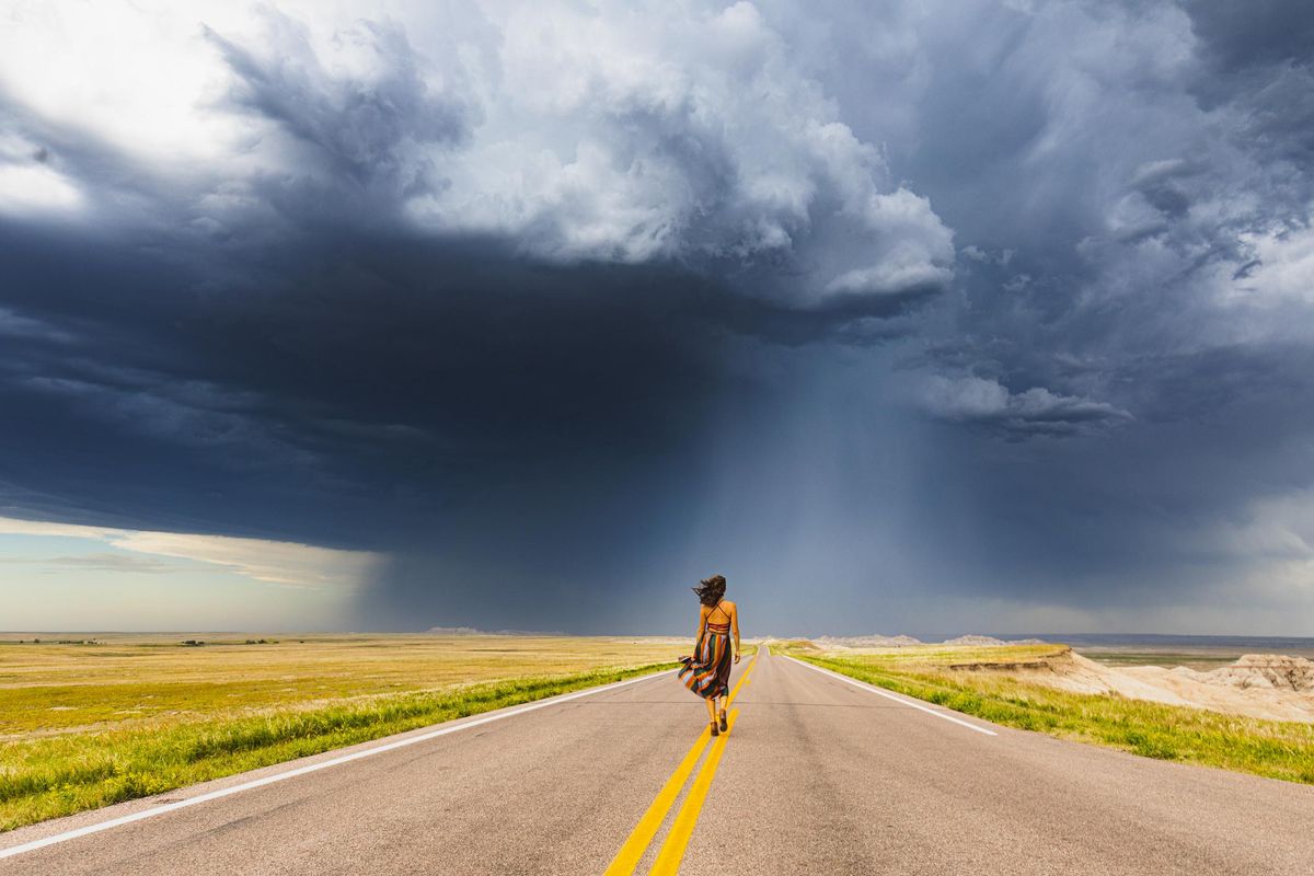 Woman in colorful dress walking down road toward dark stormy clouds during rainstorm