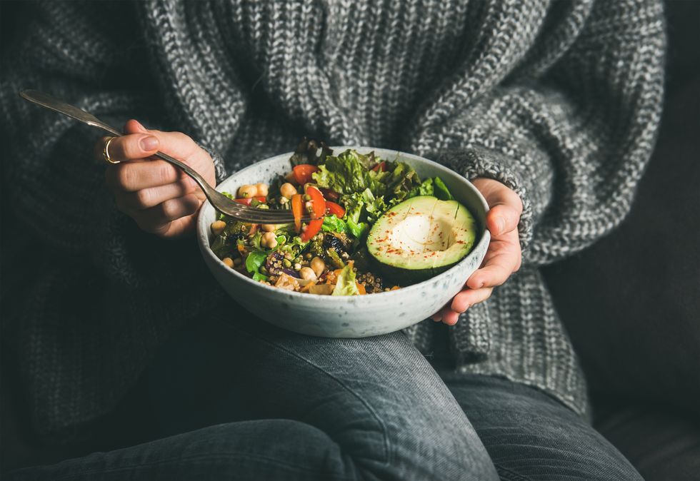 Woman in grey jeans and sweater eating fresh salad, avocado half, grains, beans, roasted vegetables from Buddha bowl