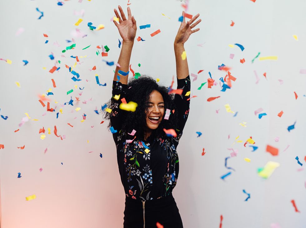 woman laughing under colorful confetti, raised her hands up