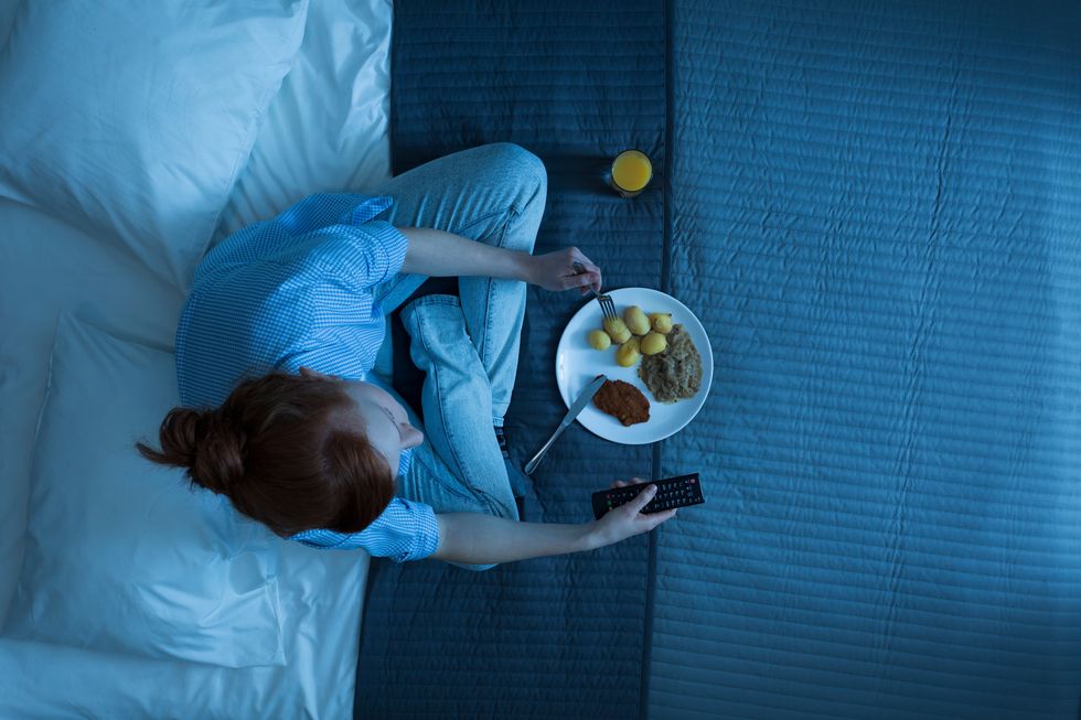 woman sitting on a bed, watching tv, eating dinner and holding a remote control