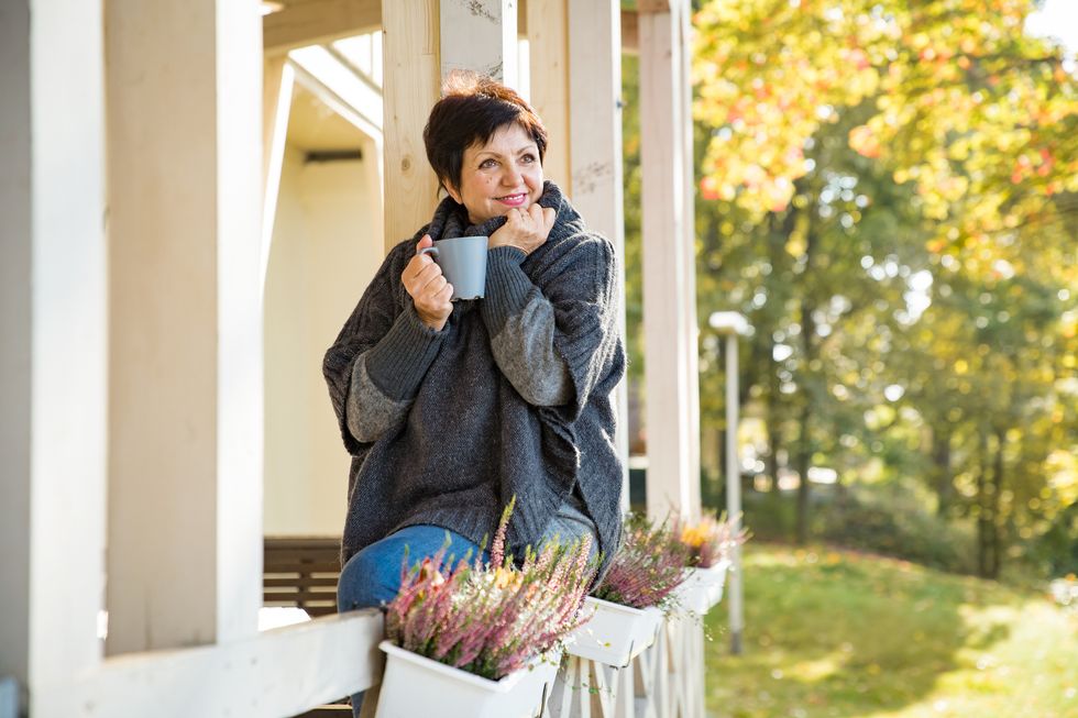 woman standing on Cozy wooden terrace with cup of hot coffee wrapped up in knitted warm sweater, happy smile