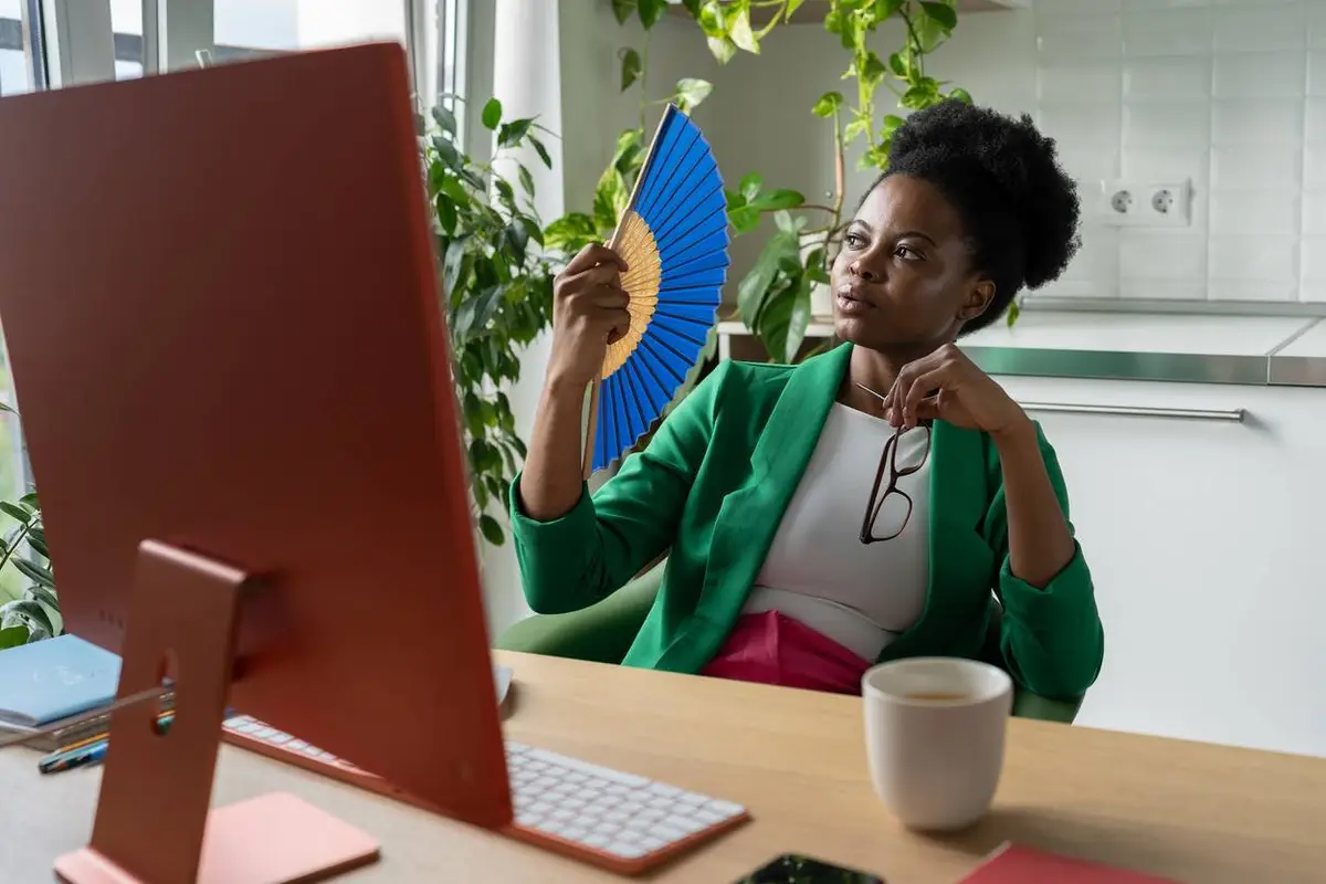 woman uses hand fan sits at desk with computer due to hot flashes