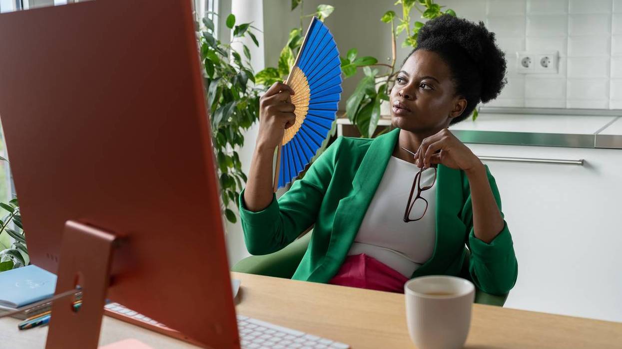 woman uses hand fan sits at desk with computer due to hot flashes