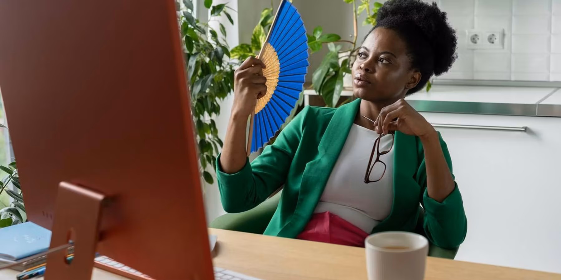 woman uses hand fan sits at desk with computer due to hot flashes