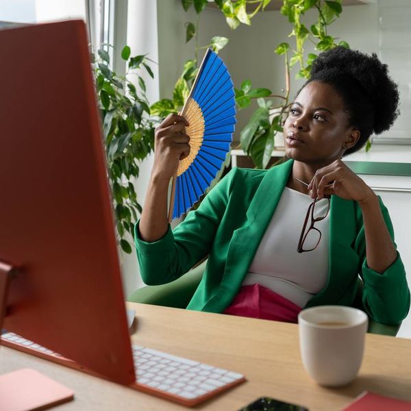woman uses hand fan sits at desk with computer due to hot flashes