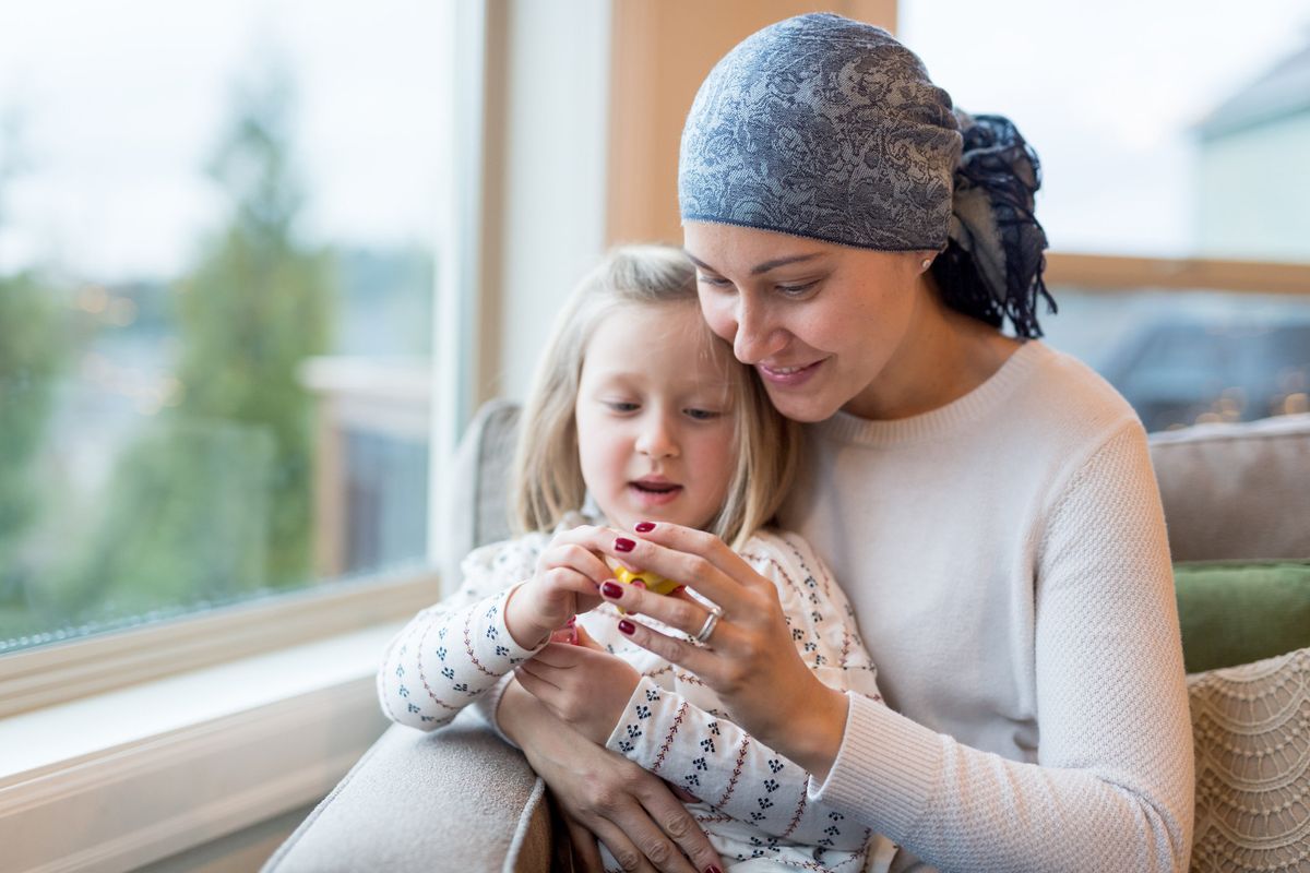 woman with cancer holds her preschool-age daughter in her lap by their living window