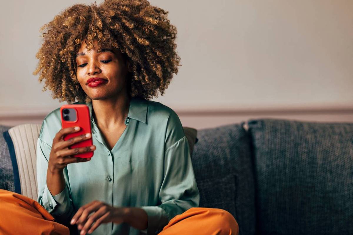 woman with curly hair browses her phone with a contented expression while sitting on a sofa