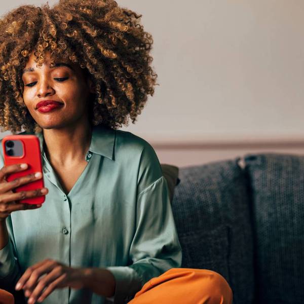woman with curly hair browses her phone with a contented expression while sitting on a sofa