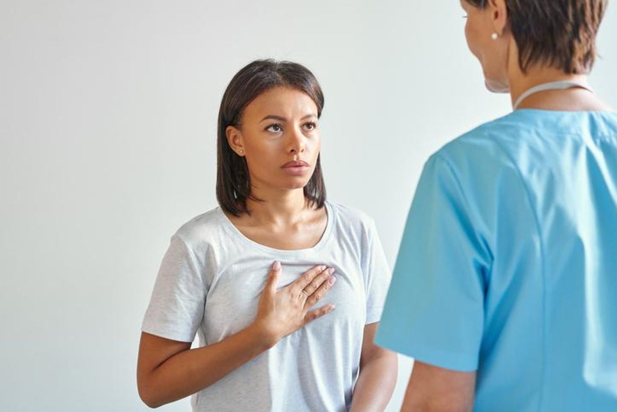 Worried young afro american woman patient touching chest, explaining symptoms and complaining about heartache to female doctor in blue uniform while visiting clinic office