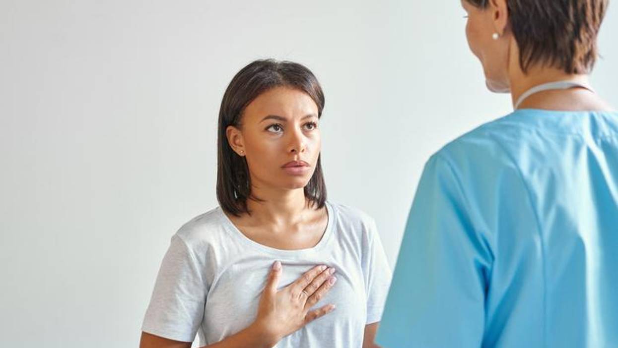 Worried young afro american woman patient touching chest, explaining symptoms and complaining about heartache to female doctor in blue uniform while visiting clinic office